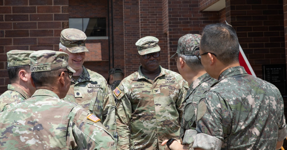 A group of military personnel in uniform are engaged in conversation outside a brick building. The group includes U.S. Army soldiers in OCP (Operational Camouflage Pattern) uniforms and what appear to be Republic of Korea (ROK) Army soldiers in their distinctive digital camouflage uniforms. The interaction appears to be a formal or semi-formal meeting between allied military forces.