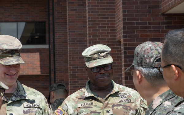 A group of military personnel in uniform are engaged in conversation outside a brick building. The group includes U.S. Army soldiers in OCP (Operational Camouflage Pattern) uniforms and what appear to be Republic of Korea (ROK) Army soldiers in their distinctive digital camouflage uniforms. The interaction appears to be a formal or semi-formal meeting between allied military forces.