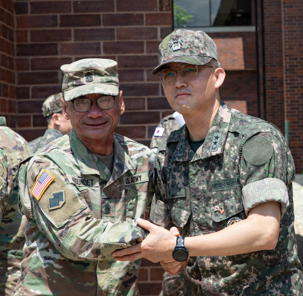 Two military personnel are posing together for a photo, shaking hands in a formal greeting. One is a U.S. Army soldier wearing OCP (Operational Camouflage Pattern) uniform with the name tape 'KOUNDET' and a Command Sergeant Major rank insignia, while the other wears a Republic of Korea (ROK) Army digital camouflage uniform with a two-star general rank insignia. The setting appears to be an outdoor area adjacent to a brick building, suggesting a bilateral military engagement or visit.