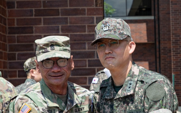 Two military personnel are posing together for a photo, shaking hands in a formal greeting. One is a U.S. Army soldier wearing OCP (Operational Camouflage Pattern) uniform with the name tape 'KOUNDET' and a Command Sergeant Major rank insignia, while the other wears a Republic of Korea (ROK) Army digital camouflage uniform with a two-star general rank insignia. The setting appears to be an outdoor area adjacent to a brick building, suggesting a bilateral military engagement or visit.