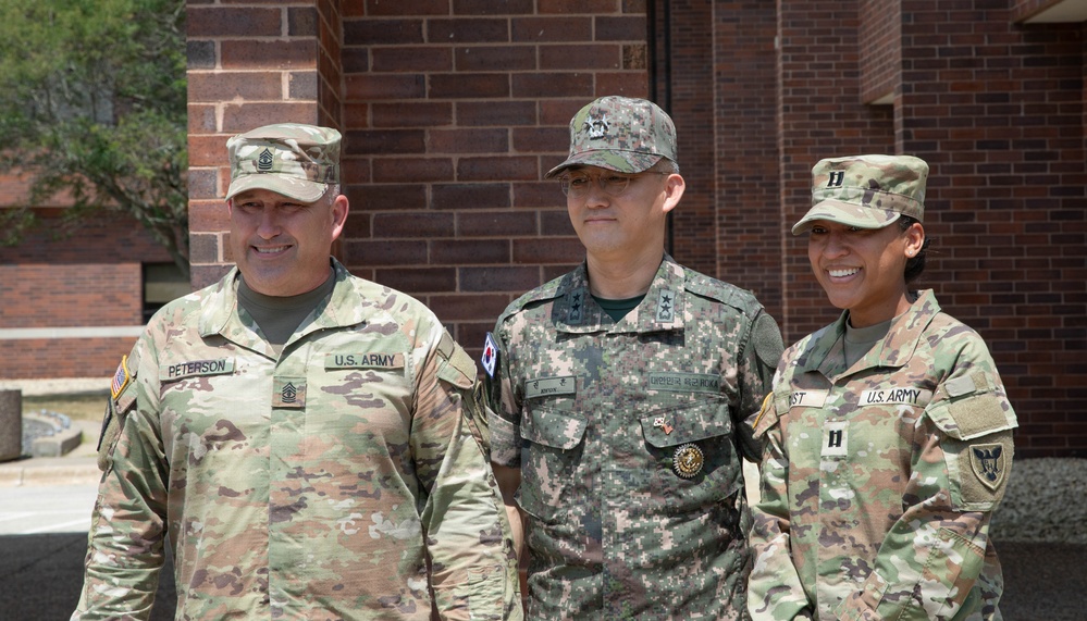 Three military personnel are posing together in front of a brick building. Two individuals wear U.S. Army OCP (Operational Camouflage Pattern) uniforms, while the center individual wears a Republic of Korea (ROK) Army digital camouflage uniform. The group appears to be participating in a joint military engagement or official visit.