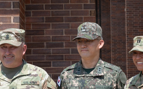 Three military personnel are posing together in front of a brick building. Two individuals wear U.S. Army OCP (Operational Camouflage Pattern) uniforms, while the center individual wears a Republic of Korea (ROK) Army digital camouflage uniform. The group appears to be participating in a joint military engagement or official visit.