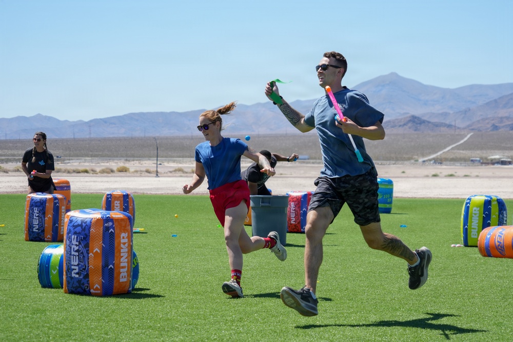 Two adults are running and playing on an artificial turf field set up as a Nerf battle arena, identifiable by branded 'NERF BUNKR' inflatable obstacles in blue and orange. The setting appears to be an outdoor desert environment, likely in the American Southwest based on the arid landscape and mountains visible in the background. Participants are holding colorful Nerf blasters and appear to be mid-game.