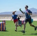 Two adults are running and playing on an artificial turf field set up as a Nerf battle arena, identifiable by branded 'NERF BUNKR' inflatable obstacles in blue and orange. The setting appears to be an outdoor desert environment, likely in the American Southwest based on the arid landscape and mountains visible in the background. Participants are holding colorful Nerf blasters and appear to be mid-game.