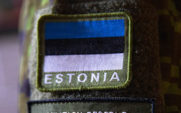 Close-up photograph of two military patches on a camouflage uniform sleeve. The upper patch displays the Estonian national flag (blue, black, and white horizontal stripes) with the word 'ESTONIA' embroidered below it. The lower patch represents the Military Reserve Exchange Program (MREP), featuring stylized lettering incorporating both the American flag and Estonian flag color schemes.