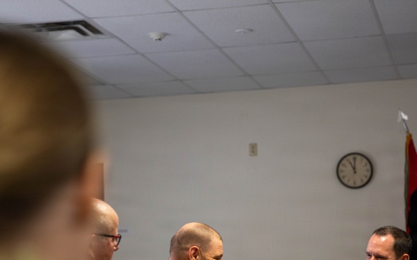 A military meeting or briefing is taking place in a conference room, with personnel from what appear to be at least two different nations present, identifiable by differing camouflage uniform patterns. The individuals seated at the table appear engaged in conversation, with relaxed and smiling expressions suggesting a collegial atmosphere. An American flag and what appears to be a unit or organizational flag are visible in the background.