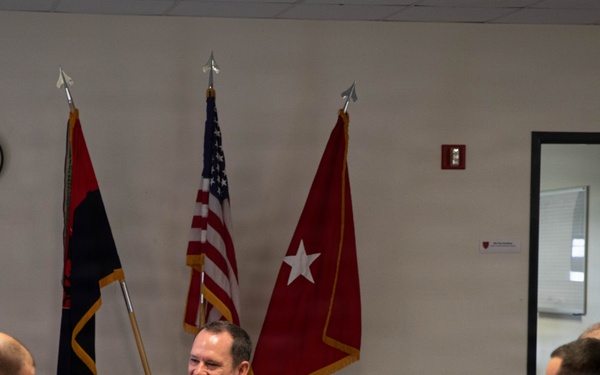 A group of military personnel from at least two different nations are seated around a conference table in what appears to be a briefing or meeting room. A U.S. Army officer in OCP (Operational Camouflage Pattern) uniform sits centrally, smiling, flanked by personnel wearing a distinctive green camouflage pattern consistent with Canadian Armed Forces uniforms. Three flags are displayed behind the central figure, including the U.S. flag and what appears to be a U.S. Army general officer's flag.