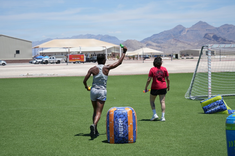 Two individuals are playing a Nerf-branded outdoor game on an artificial turf field in what appears to be a desert location, likely Nevada based on the mountain backdrop. One player in a gray tank top raises a green object (likely a Nerf gun or flag) while the other in a red shirt stands near a soccer goal. Inflatable Nerf-branded obstacles are positioned on the field as cover.