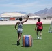 Two individuals are playing a Nerf-branded outdoor game on an artificial turf field in what appears to be a desert location, likely Nevada based on the mountain backdrop. One player in a gray tank top raises a green object (likely a Nerf gun or flag) while the other in a red shirt stands near a soccer goal. Inflatable Nerf-branded obstacles are positioned on the field as cover.