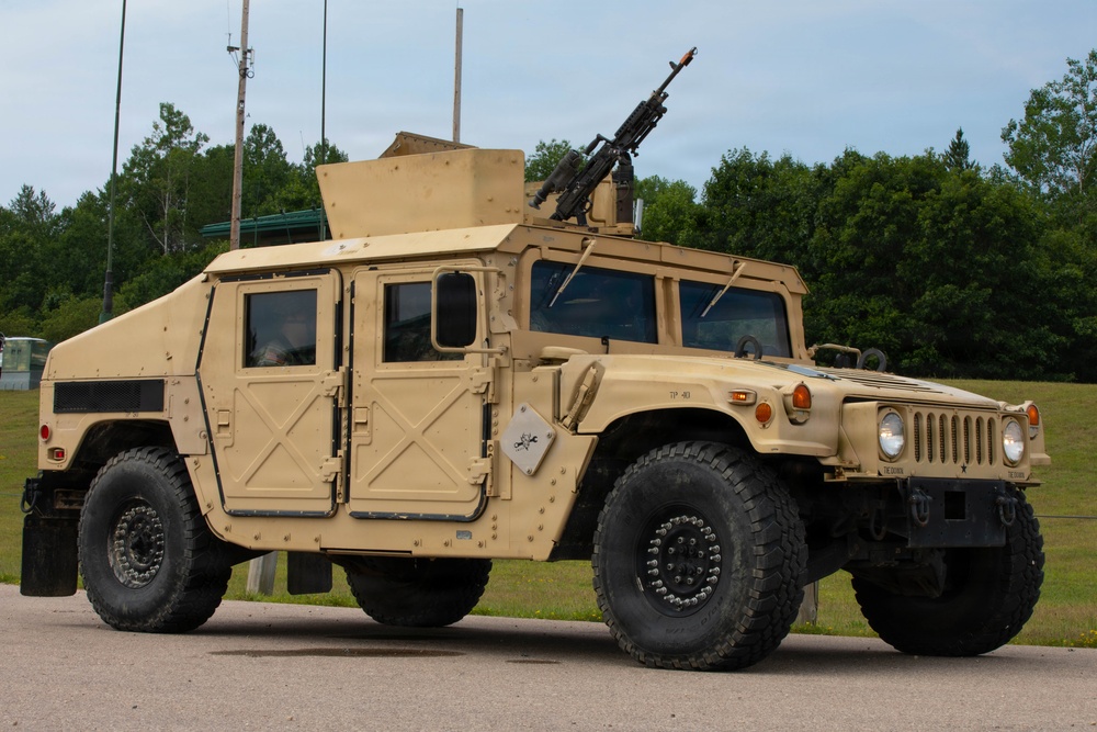 A tan/desert-colored military HMMWV (High Mobility Multipurpose Wheeled Vehicle), commonly known as a Humvee, is parked on a paved surface in what appears to be a military installation or training area. The vehicle is equipped with an armored body kit and a crew-served weapon mounted on the roof turret. The setting includes green trees, grass, and communication poles in the background under an overcast sky.