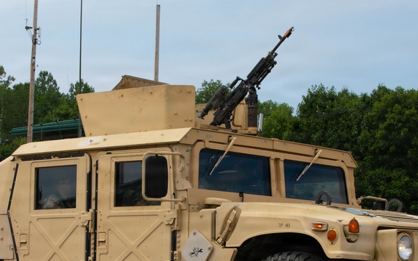 A tan/desert-colored military HMMWV (High Mobility Multipurpose Wheeled Vehicle), commonly known as a Humvee, is parked on a paved surface in what appears to be a military installation or training area. The vehicle is equipped with an armored body kit and a crew-served weapon mounted on the roof turret. The setting includes green trees, grass, and communication poles in the background under an overcast sky.