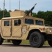A tan/desert-colored military HMMWV (High Mobility Multipurpose Wheeled Vehicle), commonly known as a Humvee, is parked on a paved surface in what appears to be a military installation or training area. The vehicle is equipped with an armored body kit and a crew-served weapon mounted on the roof turret. The setting includes green trees, grass, and communication poles in the background under an overcast sky.
