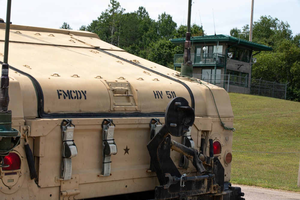 The image shows the rear section of a military vehicle painted in desert tan/beige camouflage, stenciled with the markings 'FMCDY' and 'HV 511' along with a military star emblem. The vehicle appears to be a large tactical military truck or launcher system, equipped with tow hooks, straps, and antenna components. In the background, a guard tower or observation post with a green roof is visible, surrounded by chain-link fencing and tree line.