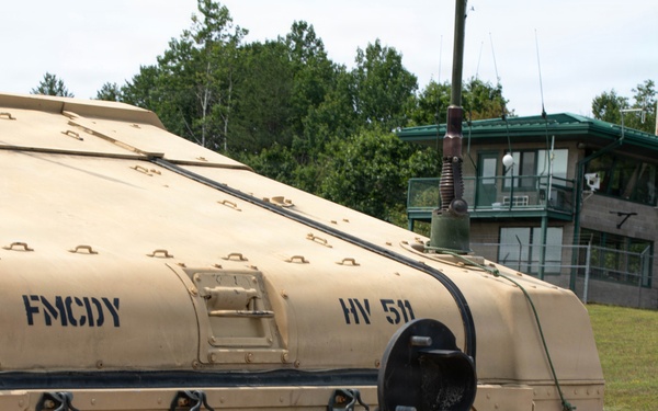 The image shows the rear section of a military vehicle painted in desert tan/beige camouflage, stenciled with the markings 'FMCDY' and 'HV 511' along with a military star emblem. The vehicle appears to be a large tactical military truck or launcher system, equipped with tow hooks, straps, and antenna components. In the background, a guard tower or observation post with a green roof is visible, surrounded by chain-link fencing and tree line.