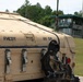 The image shows the rear section of a military vehicle painted in desert tan/beige camouflage, stenciled with the markings 'FMCDY' and 'HV 511' along with a military star emblem. The vehicle appears to be a large tactical military truck or launcher system, equipped with tow hooks, straps, and antenna components. In the background, a guard tower or observation post with a green roof is visible, surrounded by chain-link fencing and tree line.