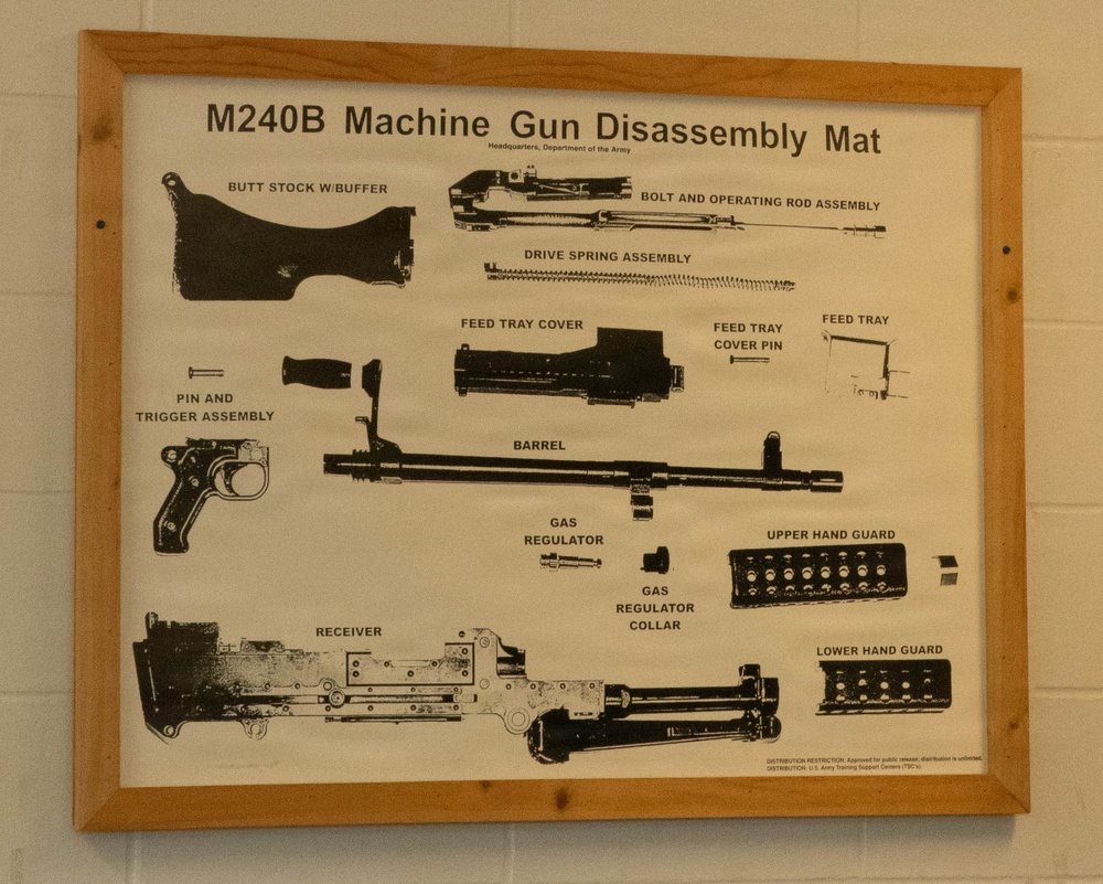 A framed instructional poster mounted on a white cinder block wall displaying the disassembly diagram of an M240B Machine Gun. The poster is issued by Headquarters, Department of the Army, and shows all major components laid out and labeled. It is presented as a training aid, likely displayed in a military facility.