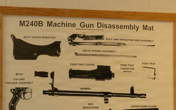 A framed instructional poster mounted on a white cinder block wall displaying the disassembly diagram of an M240B Machine Gun. The poster is issued by Headquarters, Department of the Army, and shows all major components laid out and labeled. It is presented as a training aid, likely displayed in a military facility.