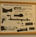 A framed instructional poster mounted on a white cinder block wall displaying the disassembly diagram of an M240B Machine Gun. The poster is issued by Headquarters, Department of the Army, and shows all major components laid out and labeled. It is presented as a training aid, likely displayed in a military facility.