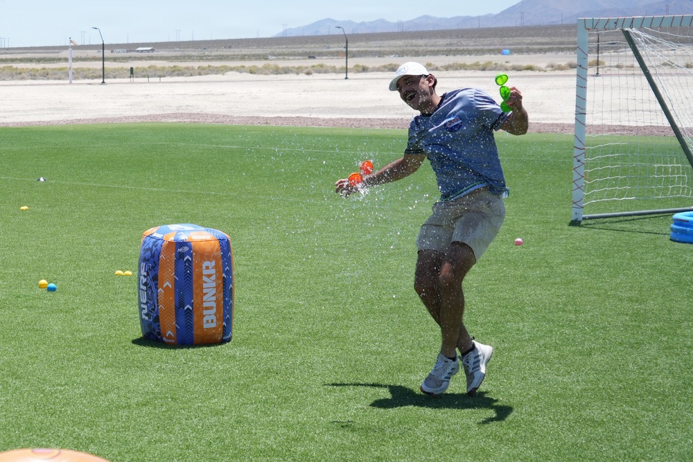 A man is playing an outdoor water balloon game on an artificial turf field in what appears to be a desert environment. He is caught mid-action as a water balloon bursts in his hands, soaking him with water. The setting includes Nerf-branded inflatable bunkers and a soccer goal, suggesting an organized outdoor recreational event.