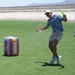 A man is playing an outdoor water balloon game on an artificial turf field in what appears to be a desert environment. He is caught mid-action as a water balloon bursts in his hands, soaking him with water. The setting includes Nerf-branded inflatable bunkers and a soccer goal, suggesting an organized outdoor recreational event.