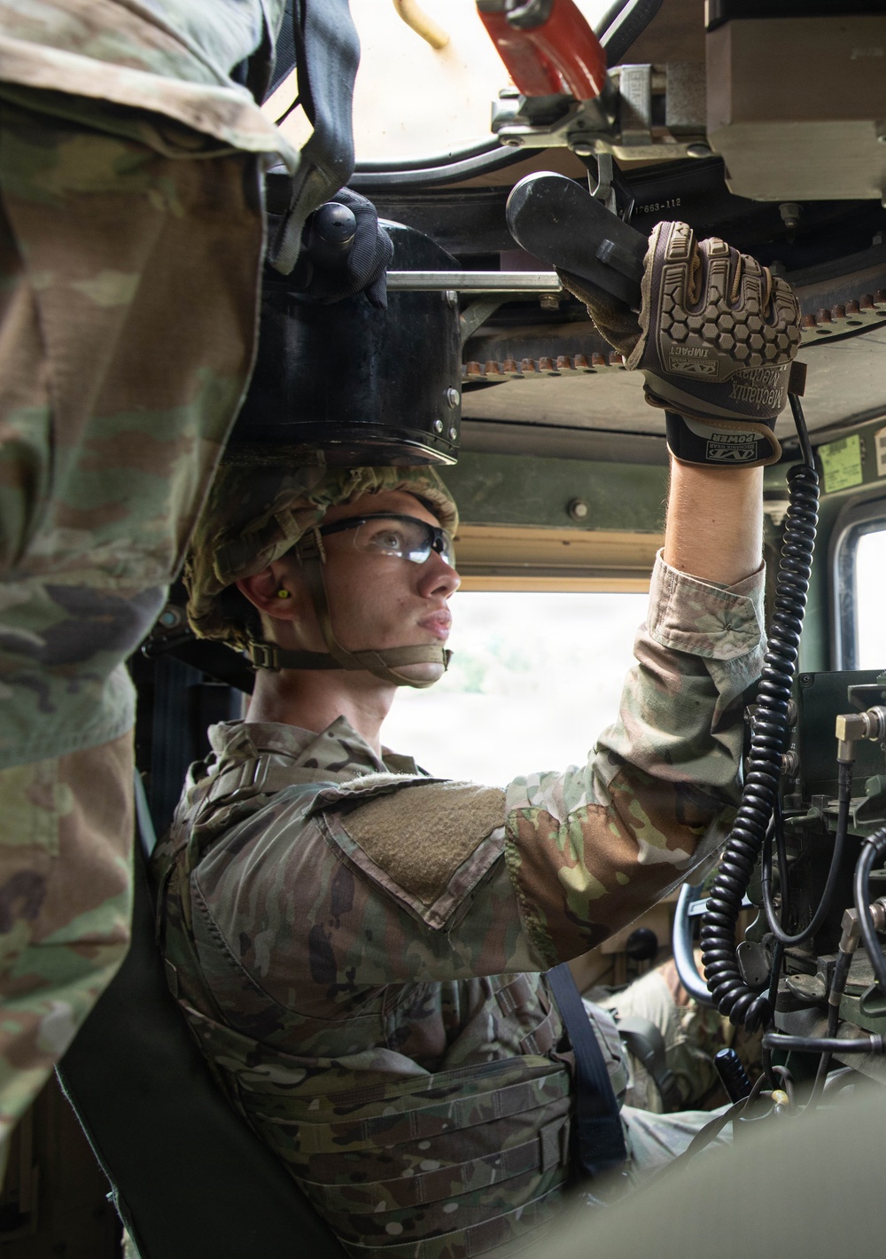 A U.S. Army soldier in OCP (Operational Camouflage Pattern) uniform is working inside a military vehicle, likely a HMMWV (Humvee). The soldier is wearing Mechanix Impact gloves, protective eyewear, a combat helmet, and body armor while manipulating overhead mechanical components. A second soldier is partially visible in the background, seated in the driver's position.