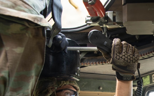 A U.S. Army soldier in OCP (Operational Camouflage Pattern) uniform is working inside a military vehicle, likely a HMMWV (Humvee). The soldier is wearing Mechanix Impact gloves, protective eyewear, a combat helmet, and body armor while manipulating overhead mechanical components. A second soldier is partially visible in the background, seated in the driver's position.