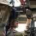 A U.S. Army soldier in OCP (Operational Camouflage Pattern) uniform is working inside a military vehicle, likely a HMMWV (Humvee). The soldier is wearing Mechanix Impact gloves, protective eyewear, a combat helmet, and body armor while manipulating overhead mechanical components. A second soldier is partially visible in the background, seated in the driver's position.