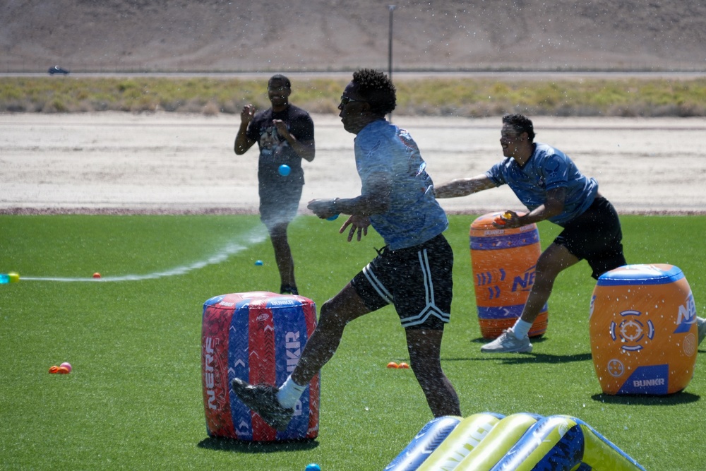 Three individuals are engaged in an outdoor water battle game on an artificial turf field using Nerf/Bunkr branded inflatable barriers. Water is visibly spraying through the air as participants dodge and maneuver around colorful inflatable obstacles. The setting appears to be a sunny, arid environment consistent with a desert climate.