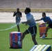 Three individuals are engaged in an outdoor water battle game on an artificial turf field using Nerf/Bunkr branded inflatable barriers. Water is visibly spraying through the air as participants dodge and maneuver around colorful inflatable obstacles. The setting appears to be a sunny, arid environment consistent with a desert climate.