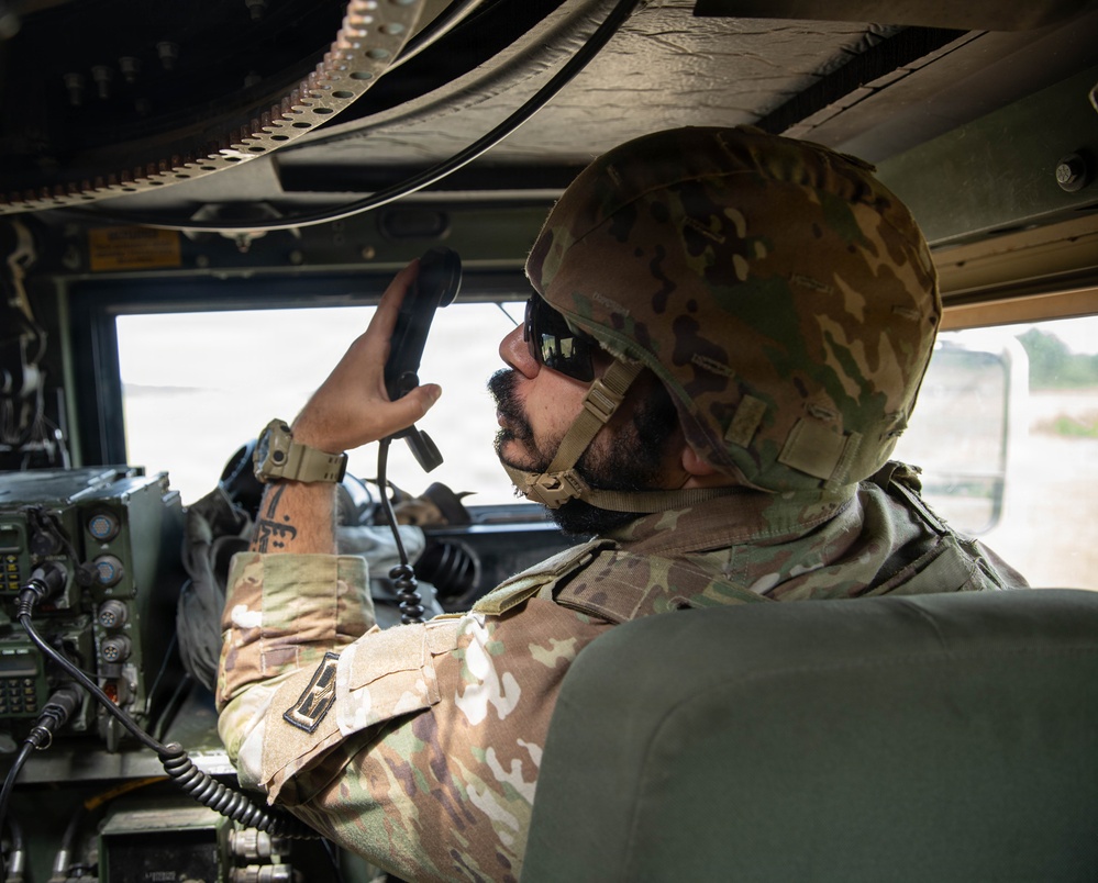 A military service member in OCP (Operational Camouflage Pattern) uniform is seated inside what appears to be a military vehicle, likely a HMMWV (Humvee). The soldier is holding a radio handset to their mouth, actively communicating, while surrounded by military radio equipment and communications gear. The interior setting and equipment suggest a tactical communications or command role.