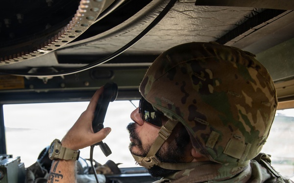 A military service member in OCP (Operational Camouflage Pattern) uniform is seated inside what appears to be a military vehicle, likely a HMMWV (Humvee). The soldier is holding a radio handset to their mouth, actively communicating, while surrounded by military radio equipment and communications gear. The interior setting and equipment suggest a tactical communications or command role.