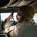 A military service member in OCP (Operational Camouflage Pattern) uniform is seated inside what appears to be a military vehicle, likely a HMMWV (Humvee). The soldier is holding a radio handset to their mouth, actively communicating, while surrounded by military radio equipment and communications gear. The interior setting and equipment suggest a tactical communications or command role.