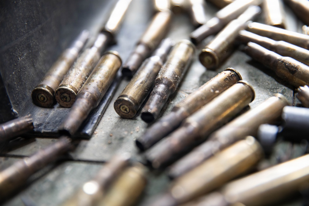 The image shows a collection of spent brass cartridge casings scattered on a metal surface. The casings appear used and show signs of oxidation, dirt, and deformation at the open ends. Some casings are partially in focus while others are blurred due to shallow depth of field.