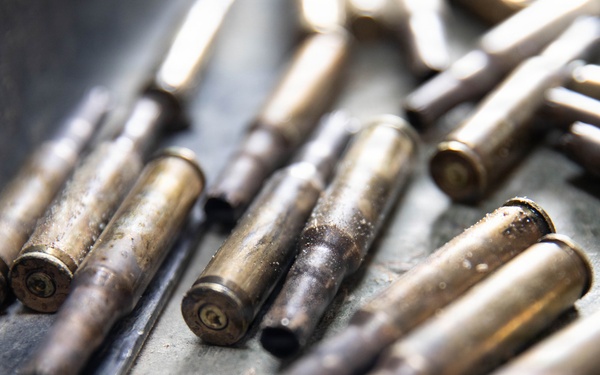 The image shows a collection of spent brass cartridge casings scattered on a metal surface. The casings appear used and show signs of oxidation, dirt, and deformation at the open ends. Some casings are partially in focus while others are blurred due to shallow depth of field.