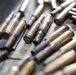 The image shows a collection of spent brass cartridge casings scattered on a metal surface. The casings appear used and show signs of oxidation, dirt, and deformation at the open ends. Some casings are partially in focus while others are blurred due to shallow depth of field.
