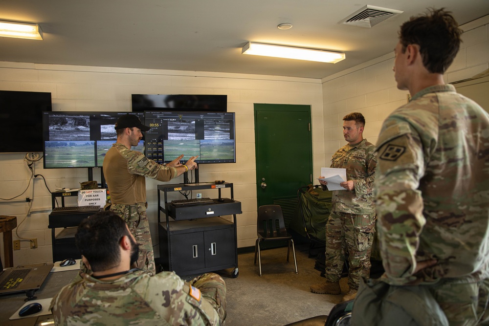 Military personnel in MultiCam/OCP camouflage uniforms are gathered in what appears to be a tactical operations or after-action review (AAR) room. One soldier is pointing at multiple monitors displaying what appear to be surveillance or drone feed imagery, while others observe and take notes. A sign visible on the desk references 'AAR PURPOSES ONLY.'