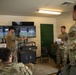 Military personnel in MultiCam/OCP camouflage uniforms are gathered in what appears to be a tactical operations or after-action review (AAR) room. One soldier is pointing at multiple monitors displaying what appear to be surveillance or drone feed imagery, while others observe and take notes. A sign visible on the desk references 'AAR PURPOSES ONLY.'
