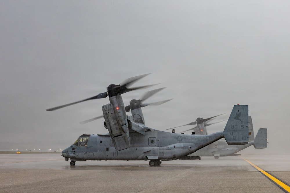 A U.S. Marines MV-22 Osprey tiltrotor aircraft sits on a wet tarmac with its rotors spinning, indicated by motion blur on the blades. The aircraft bears the tail number '8015' and Marine Corps markings, and appears to be preparing for departure or has just landed in rainy, overcast conditions. A second Osprey is partially visible in the background.