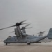 A U.S. Marines MV-22 Osprey tiltrotor aircraft sits on a wet tarmac with its rotors spinning, indicated by motion blur on the blades. The aircraft bears the tail number '8015' and Marine Corps markings, and appears to be preparing for departure or has just landed in rainy, overcast conditions. A second Osprey is partially visible in the background.