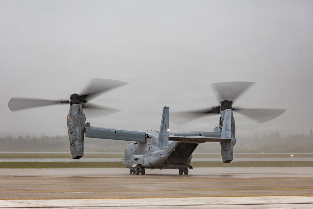 A military tiltrotor aircraft, identifiable as a Bell Boeing V-22 Osprey, is photographed on a wet tarmac during rainy, overcast conditions. The rotors are spinning, indicated by motion blur on both proprotor assemblies, suggesting the aircraft is either preparing for takeoff or has just landed. The gray, misty weather conditions reduce visibility in the background.