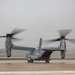 A military tiltrotor aircraft, identifiable as a Bell Boeing V-22 Osprey, is photographed on a wet tarmac during rainy, overcast conditions. The rotors are spinning, indicated by motion blur on both proprotor assemblies, suggesting the aircraft is either preparing for takeoff or has just landed. The gray, misty weather conditions reduce visibility in the background.