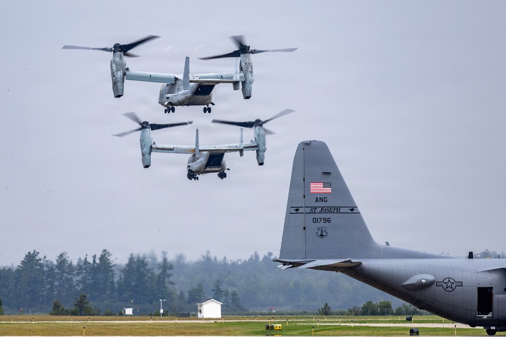 The image shows two V-22 Osprey tiltrotor aircraft in flight, appearing to be landing or hovering over a military airfield. In the foreground, the tail section of a C-130 Hercules transport aircraft is visible, marked with 'ANG' (Air National Guard), 'ST JOSEPH', and tail number '01796'. The scene appears to be at a military or Air National Guard installation, likely during a training exercise or operational mission.