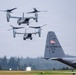 The image shows two V-22 Osprey tiltrotor aircraft in flight, appearing to be landing or hovering over a military airfield. In the foreground, the tail section of a C-130 Hercules transport aircraft is visible, marked with 'ANG' (Air National Guard), 'ST JOSEPH', and tail number '01796'. The scene appears to be at a military or Air National Guard installation, likely during a training exercise or operational mission.