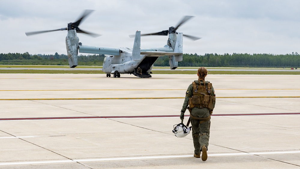 A military aviator in flight gear walks across a tarmac toward a Bell Boeing V-22 Osprey tiltrotor aircraft with its rotors spinning. The aircraft appears to be on the ground with its rear ramp open, suggesting imminent boarding or departure. The scene takes place at a military airfield surrounded by tree lines under an overcast sky.