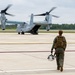 A military aviator in flight gear walks across a tarmac toward a Bell Boeing V-22 Osprey tiltrotor aircraft with its rotors spinning. The aircraft appears to be on the ground with its rear ramp open, suggesting imminent boarding or departure. The scene takes place at a military airfield surrounded by tree lines under an overcast sky.