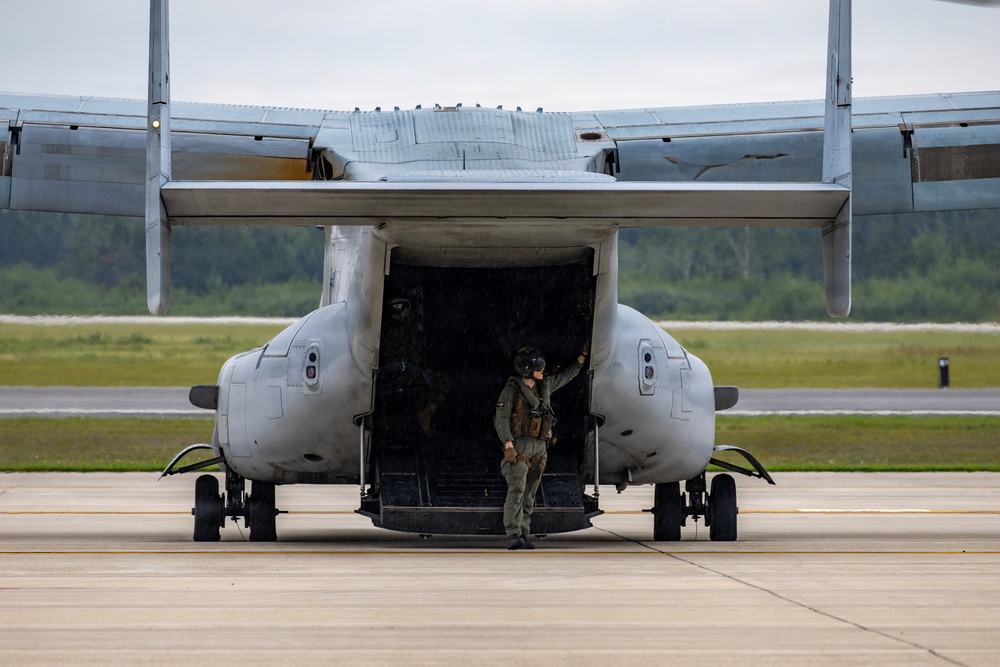 A military crew member in flight gear stands at the open rear ramp of what appears to be a Bell Boeing V-22 Osprey tiltrotor aircraft on a tarmac. The aircraft's rear cargo ramp is fully open, and the crew member appears to be conducting pre-flight or post-flight checks. The image is taken from a direct rear perspective, showing the underside of the aircraft's wing box and twin nacelles.