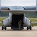 A military crew member in flight gear stands at the open rear ramp of what appears to be a Bell Boeing V-22 Osprey tiltrotor aircraft on a tarmac. The aircraft's rear cargo ramp is fully open, and the crew member appears to be conducting pre-flight or post-flight checks. The image is taken from a direct rear perspective, showing the underside of the aircraft's wing box and twin nacelles.