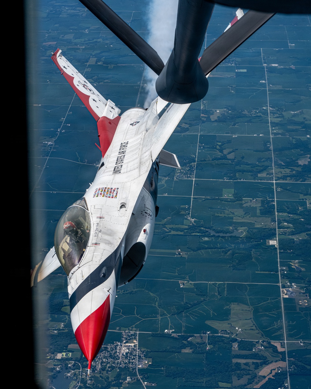 A United States Air Force Thunderbirds F-16 Fighting Falcon is captured during an aerial refueling operation with a tanker aircraft, viewed from above and behind the tanker's boom operator position. The jet displays the iconic red, white, and blue Thunderbirds livery with 'UNITED STATES AIR FORCE' lettering along the fuselage. A patchwork of agricultural fields is visible far below, suggesting the aircraft are at high altitude over a rural Midwestern landscape.