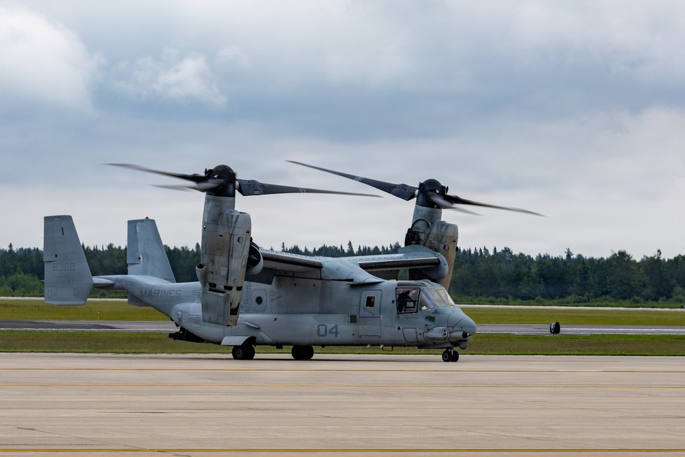 A U.S. Marines MV-22 Osprey tiltrotor aircraft is taxiing on a runway with its rotors spinning. The aircraft bears the number '04' on its fuselage and 'MARINES' markings, with a tail number partially visible as '6368'. The setting appears to be a military or joint-use airfield surrounded by coniferous trees under overcast skies.