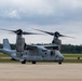 A U.S. Marines MV-22 Osprey tiltrotor aircraft is taxiing on a runway with its rotors spinning. The aircraft bears the number '04' on its fuselage and 'MARINES' markings, with a tail number partially visible as '6368'. The setting appears to be a military or joint-use airfield surrounded by coniferous trees under overcast skies.